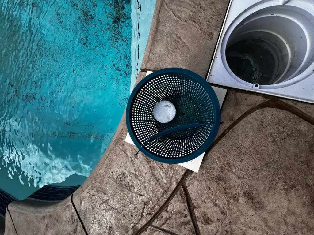 Grey Shark Pool Services in Oak Island, NC: skimmer basket with chlorine tablet by pool, blue hose on concrete near open skimmer well.