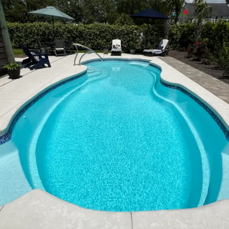 Outdoor swimming pool in Oak Island, NC, serviced by Grey Shark Pool Services; clear water, white paving, lounge chairs, and trees.