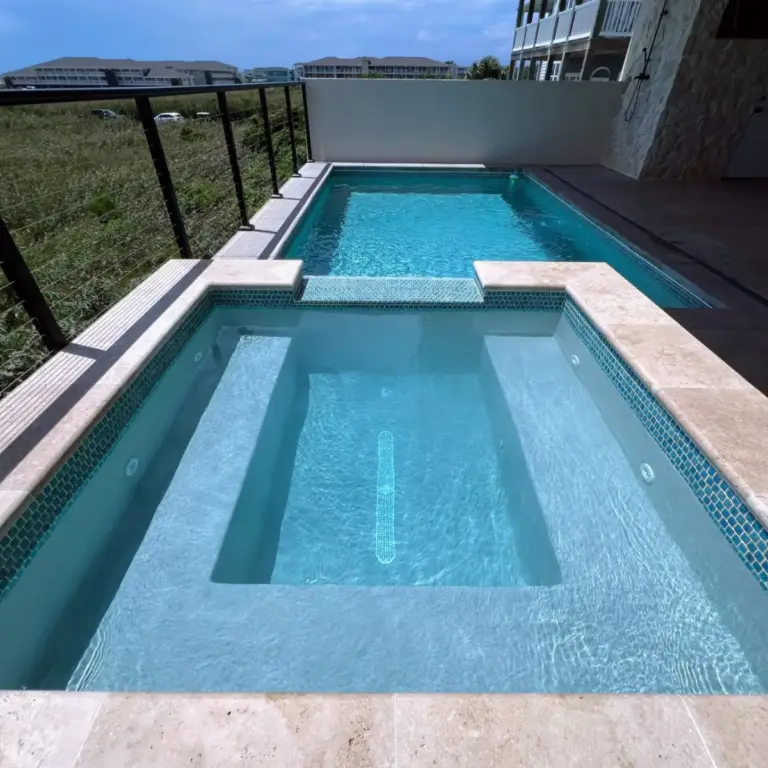 Modern outdoor pool and square hot tub serviced by Grey Shark Pool Services in Oak Island, NC, with beige tiles and metal railing.