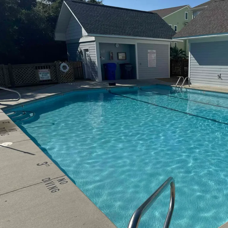 Grey Shark Pool Services in Oak Island, NC: clear outdoor pool, metal handrails, 3 FT NO DIVING sign, blue buildings, sunny sky.