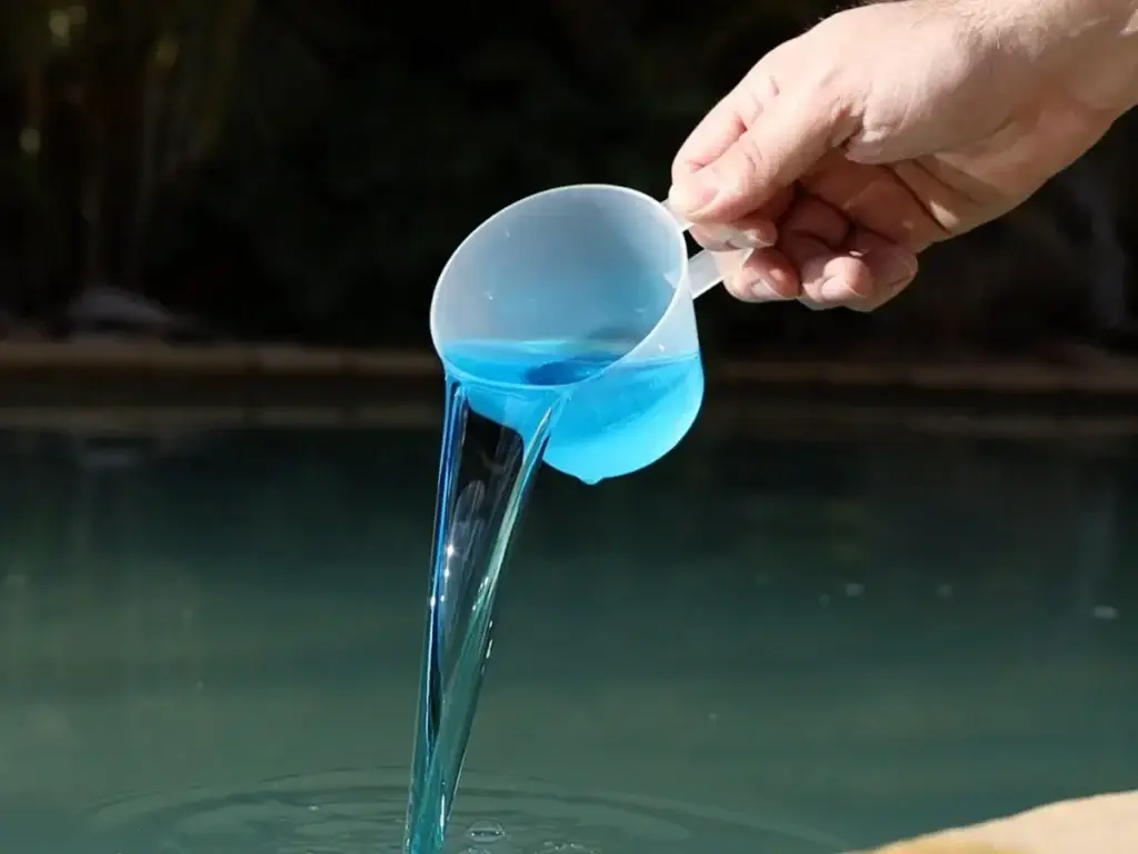 A hand pours bright blue liquid from a measuring cup into a pool, representing Grey Shark Pool Services in Oak Island, NC.